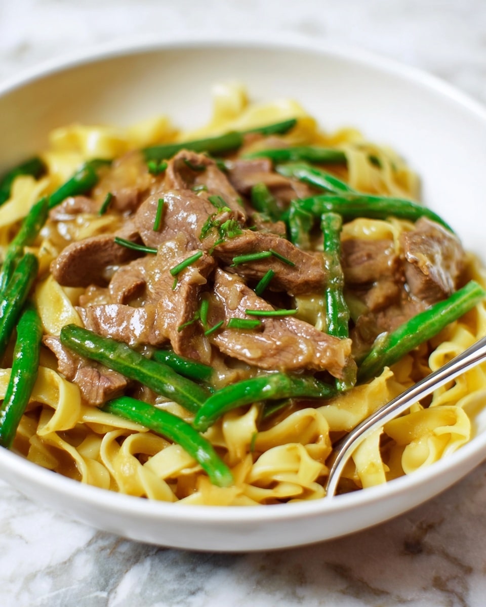 The dish is served in a white bowl filled with three main layers: the base layer consists of light yellow flat noodles arranged loosely; on top of this are green beans scattered evenly, adding bright green color and a fresh texture; the top layer contains slices of cooked beef in a light brown sauce that covers the noodles and beans lightly, giving a slightly shiny appearance; small green chopped herbs are sprinkled on top for garnish; a silver fork is placed at the bottom edge of the bowl on a white marbled surface. photo taken with an iphone --ar 4:5 --v 7