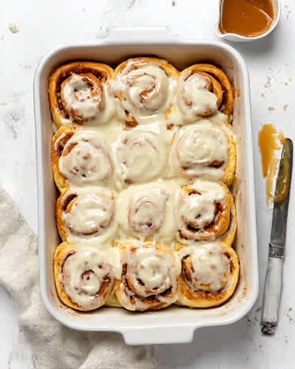 The image shows a white rectangular baking dish filled with twelve soft cinnamon rolls covered in melted white icing. The rolls are arranged in three rows of four, with golden-brown edges and a soft, fluffy texture visible. The icing is thick and creamy, generously spread over the rolls, slightly dripping into the gaps between them. The dish is placed on a white marbled surface, with a butter knife to the right side, its blade slightly touched by icing. A small portion of caramel or brown sauce is visible in a small white bowl at the top right corner. Photo taken with an iphone --ar 4:5 --v 7