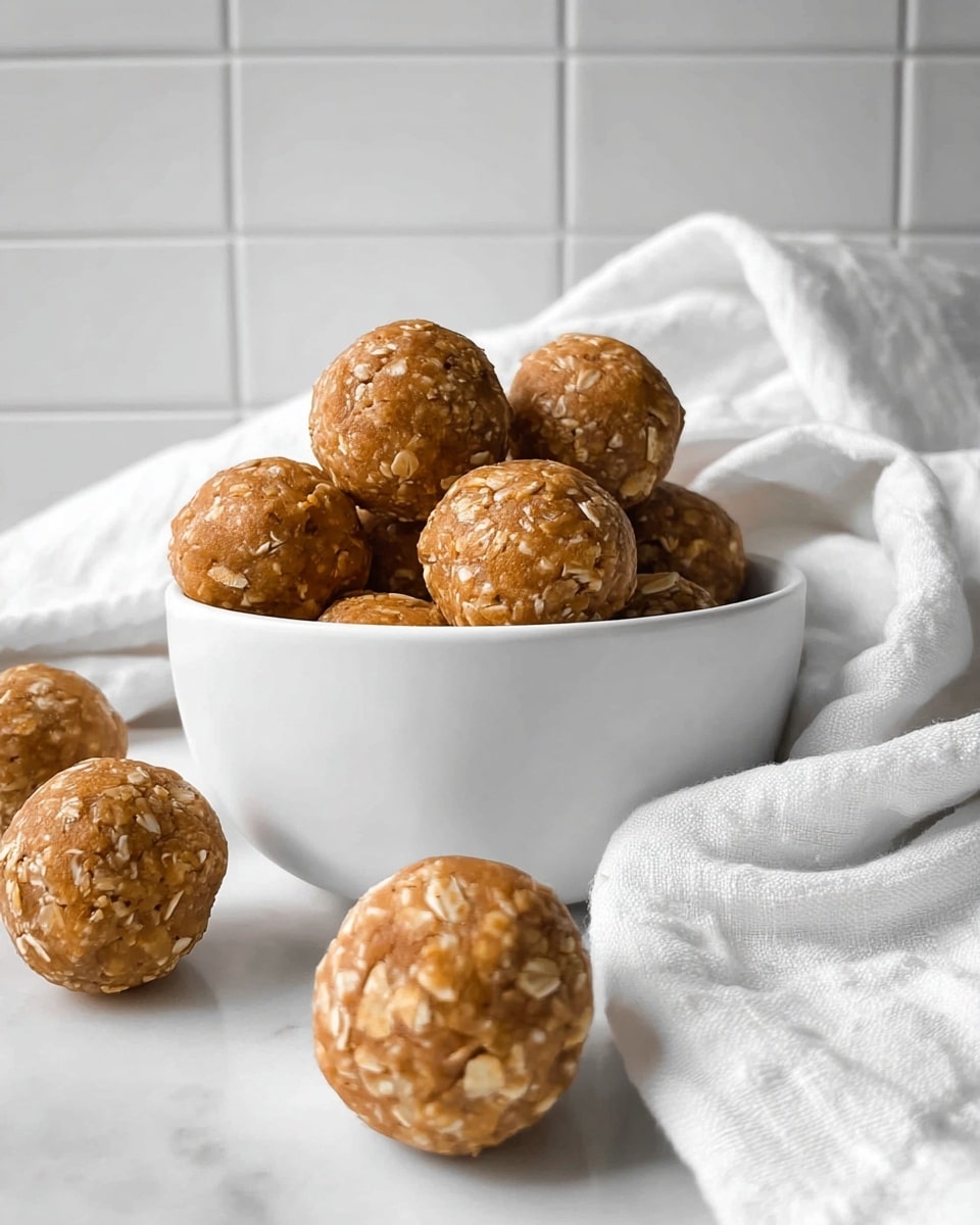 A white bowl filled with round energy balls that have a light brown color with visible oats and a slightly rough texture; several more balls are placed around the bowl on a white marbled surface, and a white cloth is softly draped in the background against a white tiled wall. photo taken with an iphone --ar 4:5 --v 7