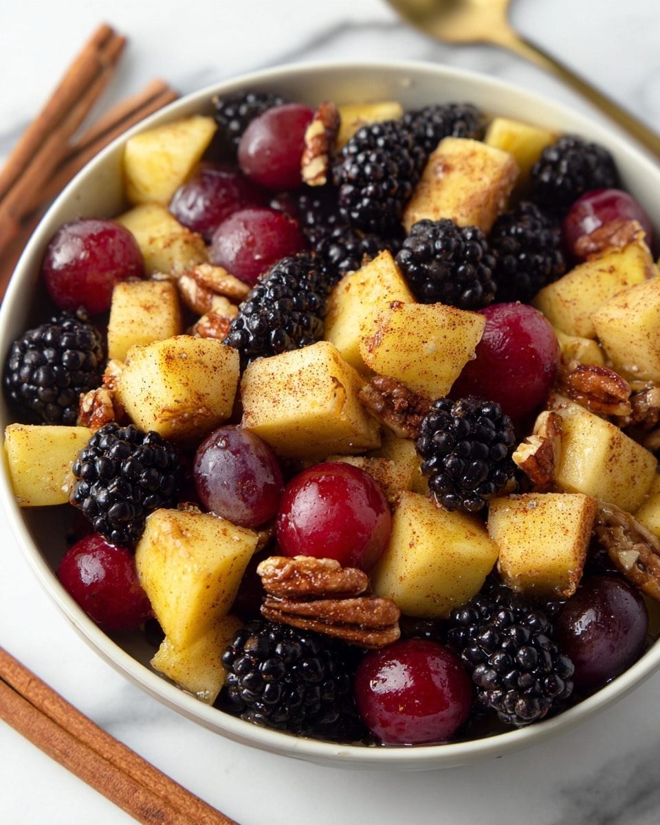 The image shows a close-up of a white bowl filled with a vibrant fruit salad. The salad contains three visible layers: large, chunky yellow apple pieces coated with a light brown spice; deep purple-black blackberries with a bumpy texture; and smooth, shiny red grapes. Scattered among the fruits are glossy, light brown pecan halves. The bowl sits on a white marbled surface with two cinnamon sticks placed nearby. The colors are rich and warm, with the textures varying from soft and juicy to crunchy. photo taken with an iphone --ar 4:5 --v 7