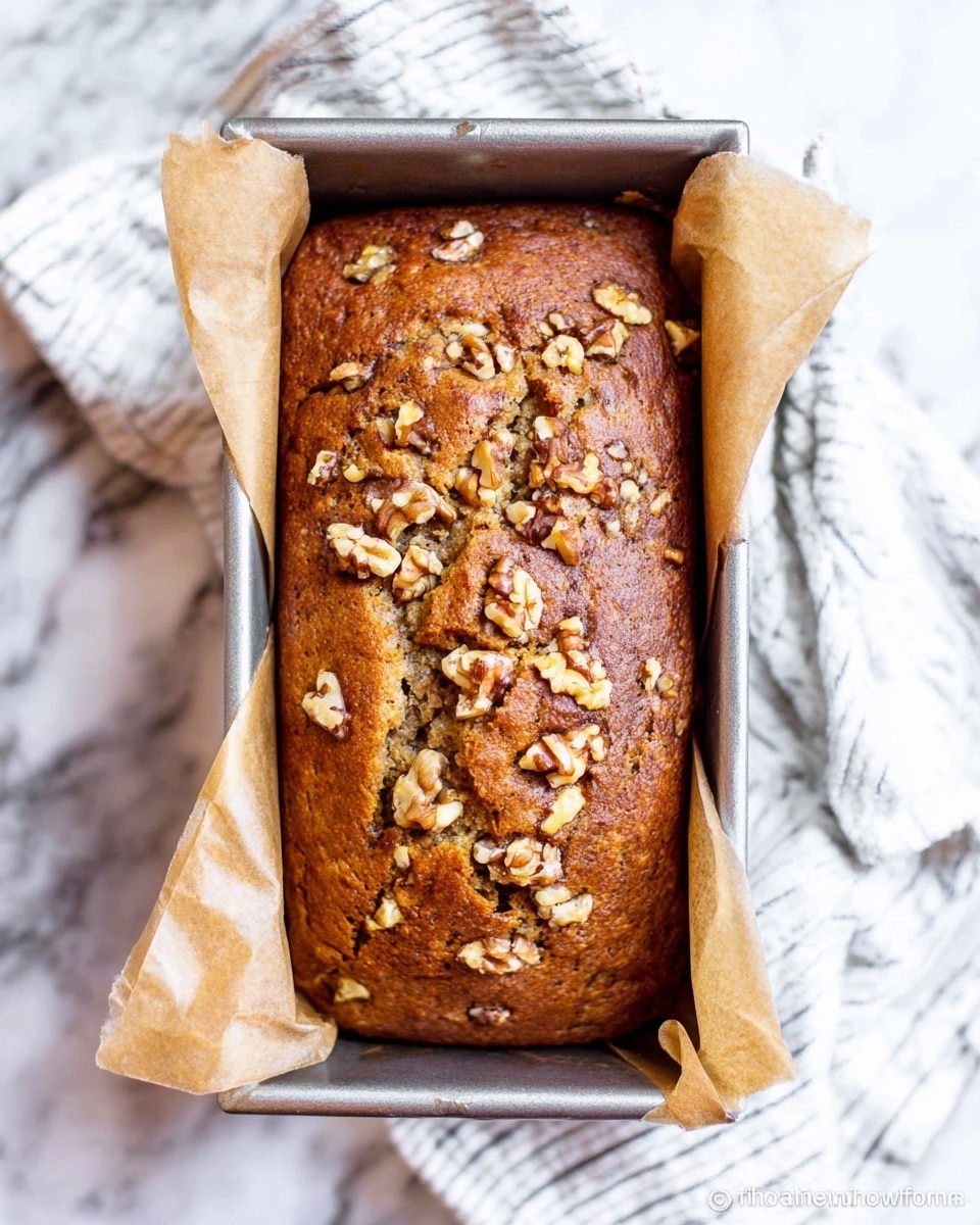 A loaf of banana bread with a golden brown, slightly cracked top covered with uneven chunks of toasted walnuts scattered all over. The bread sits snugly inside a silver metal loaf pan lined with brown parchment paper on both sides extending out. The pan rests on a white cloth with thin black stripes, placed on a white marbled surface that has subtle gray patterns. photo taken with an iphone --ar 4:5 --v 7