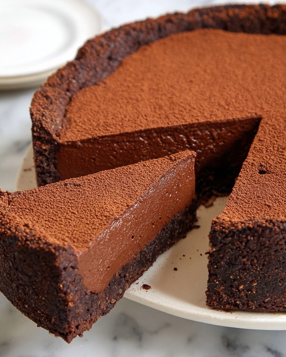 A close-up view of a single slice of rich chocolate pie placed on a white plate with slight brown speckles. The pie slice has three visible layers: a bottom thin dark brown crust with a firm texture, a thick middle layer of smooth, dense milk chocolate filling in a medium brown color, and a top layer dusted with fine cocoa powder in a darker brown shade. The edge of the pie crust is crumbly and raised with uneven texture. In the background, the remaining pie on a white plate is softly blurred. The scene sits on a white marbled texture surface. photo taken with an iphone --ar 4:5 --v 7