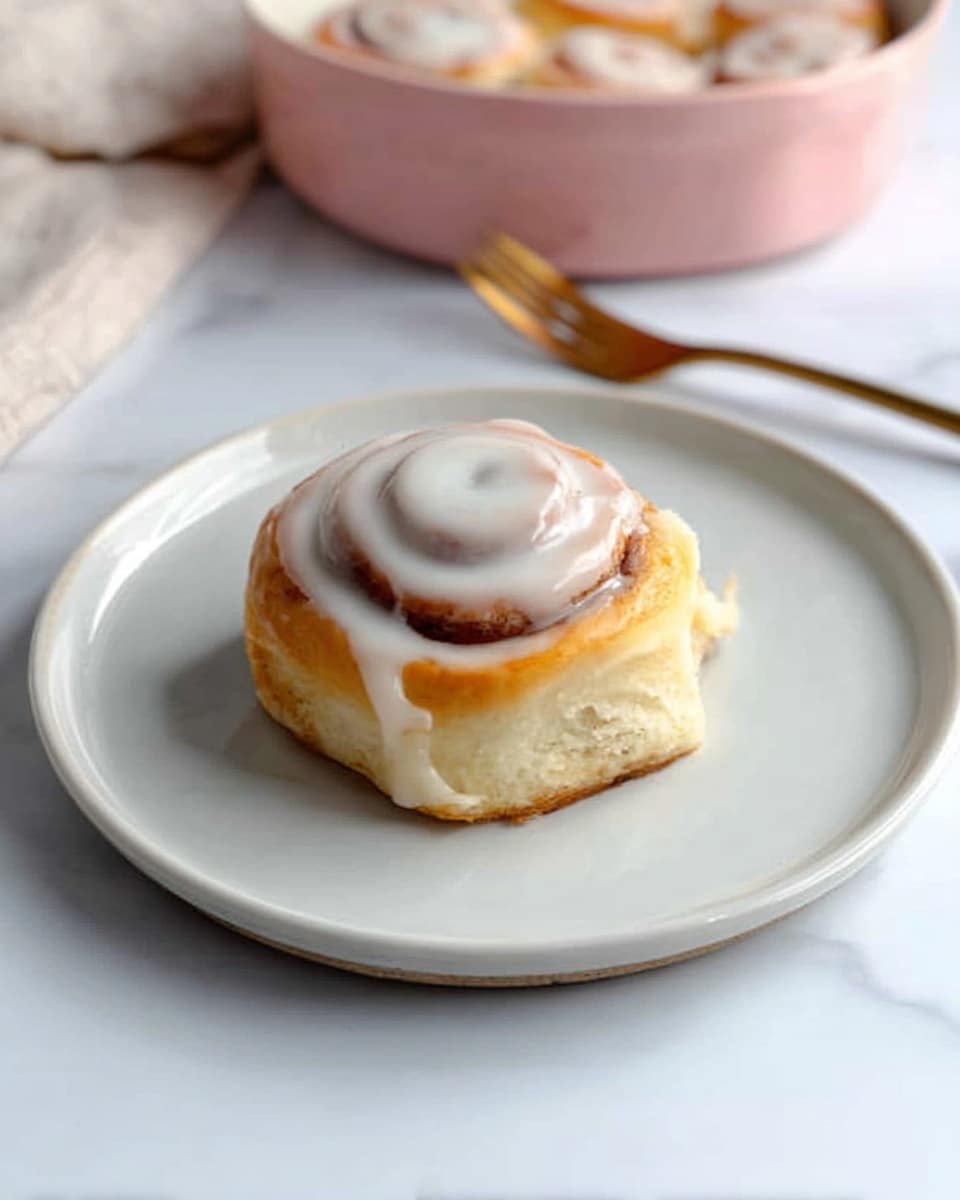 A single cinnamon roll sits in the center of a white plate on a white marbled surface. The cinnamon roll has three visible layers of soft, golden-brown dough spiraled neatly, with a thick layer of smooth, white icing covering the top and slightly dripping down the sides. Behind the plate, there is a blurred view of a white fork and a pink bowl filled with more cinnamon rolls. In the top left corner, a woman's hand is slightly reaching toward the bowl. Photo taken with an iphone --ar 4:5 --v 7
