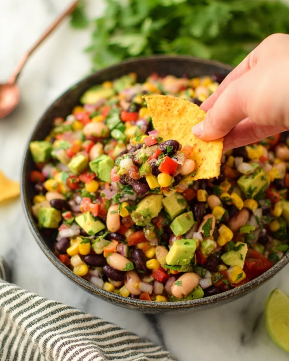 A large white bowl filled with a colorful mix of black beans, white beans, cut green avocado chunks, yellow corn, red diced bell peppers, green bell peppers, chopped red onions, tomatoes, and fresh green cilantro leaves, all mixed together giving a vibrant, fresh look. Next to the bowl on a gray striped cloth are several yellow tortilla chips. The dish is placed on a white marbled surface. photo taken with an iphone --ar 4:5 --v 7