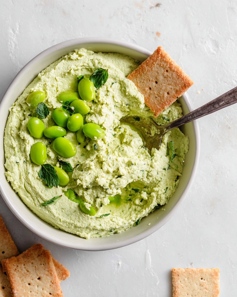 A white bowl holds a thick, creamy pale green dip with a slightly rough texture. Scattered on top are bright green edamame beans and a few green herb leaves for garnish. A silver spoon rests inside the bowl, scooping up some dip. One square whole grain cracker is partially dipped into the spread, and two more crackers lay nearby on a white marbled surface. photo taken with an iphone --ar 4:5 --v 7