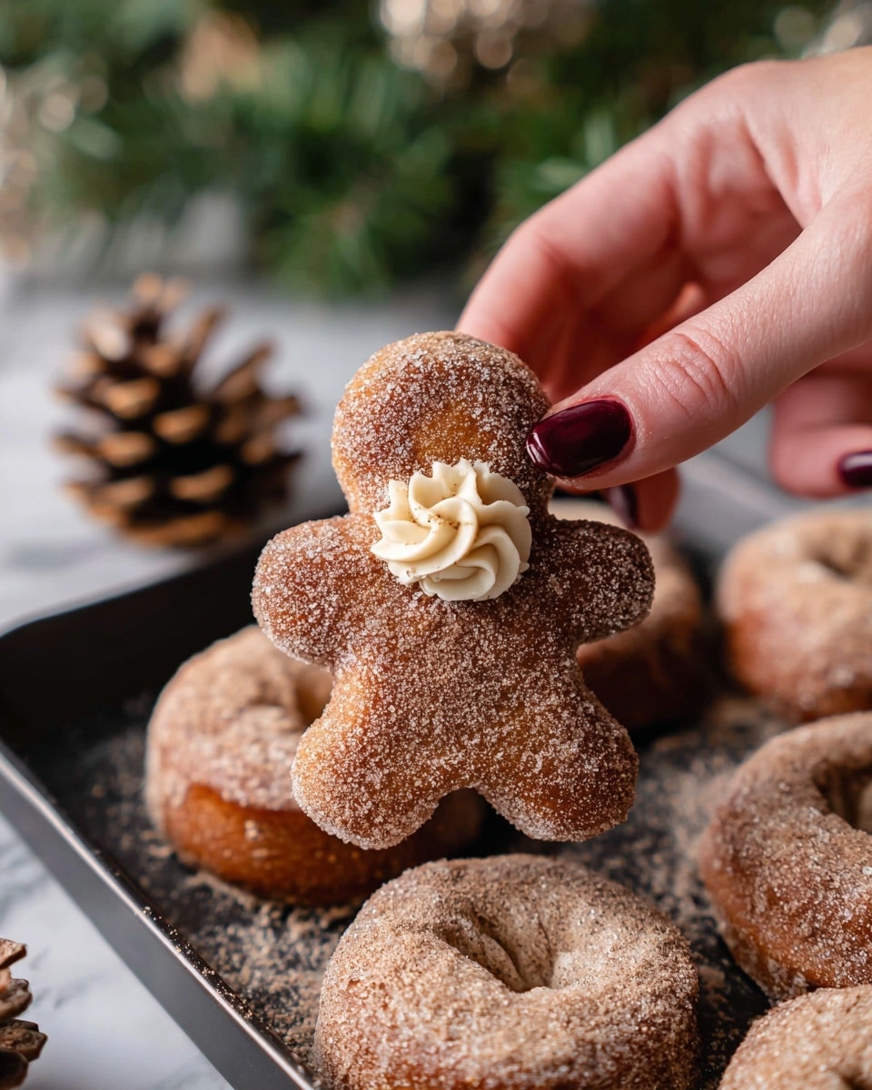 The image shows a close-up of a soft, gingerbread-shaped donut covered in a sugary cinnamon coating. The donut is shaped like a small person and has a small swirl of light beige cream with visible tiny dark specks on its chest area. The donut is being picked up delicately by a woman’s hand with dark red nail polish. The donut rests on a black tray sprinkled with extra cinnamon sugar, and other similar donuts are visible around it. The background features a blurred green pine branch and pinecone on a white marbled surface. photo taken with an iphone --ar 4:5 --v 7