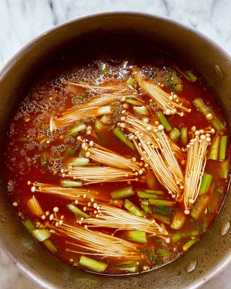 The image shows a pot filled with a rich reddish-brown broth as the base layer, clear but oily with small bubbles floating on the surface. Inside the broth, there are several bundles of light beige enoki mushrooms with thin stems and small caps, spread evenly around the pot. Bright green pieces of what looks like scallion or leek are scattered throughout, adding a fresh contrast in color. The texture of the mushrooms appears soft and delicate while the broth looks thick and steaming. The woman’s hand is not visible in this image. The pot edges are visible with a smooth surface, and the background is a white marbled texture. photo taken with an iphone --ar 4:5 --v 7