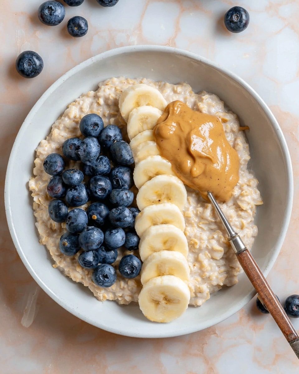 A shallow white bowl contains a creamy oatmeal base with a light beige color and soft texture, spread evenly across the bowl. On top of the oatmeal, there is a thick dollop of smooth peanut butter placed slightly off-center, with a rich golden-brown color. To the left of the peanut butter, a cluster of fresh, plump blueberries with a deep blue-purple hue sits neatly. To the right, a row of evenly sliced banana pieces, each pale yellow with a soft texture and subtle brown specks at the center, is arranged carefully. A silver spoon with a wooden handle rests inside the bowl on the left side, holding some blueberries and oatmeal. The bowl is set on a white marbled textured surface with a few scattered blueberries nearby. Photo taken with an iphone --ar 4:5 --v 7