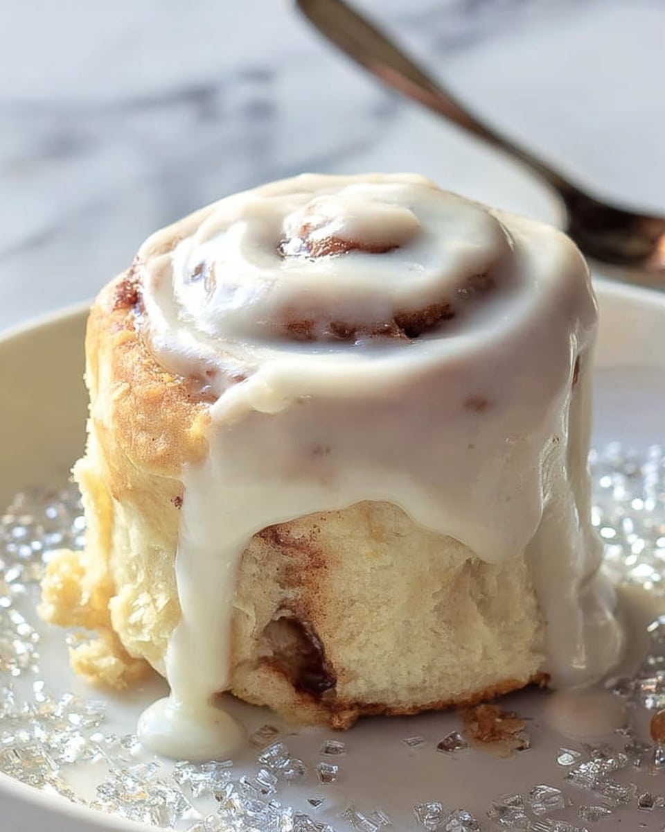 A single cinnamon roll stands tall on a white plate with sparkling sugar crystals scattered around. The roll has a soft, fluffy dough base that is light golden brown, with a visible inner swirl showing the cinnamon and sugar filling in a warm brown color. A thick layer of creamy white icing covers the top, slowly dripping down the sides, pooling slightly at the base. Next to the cinnamon roll is a shiny silver spoon partially visible. The background features a white marbled texture that adds a clean, bright feel to the image. Photo taken with an iphone --ar 4:5 --v 7