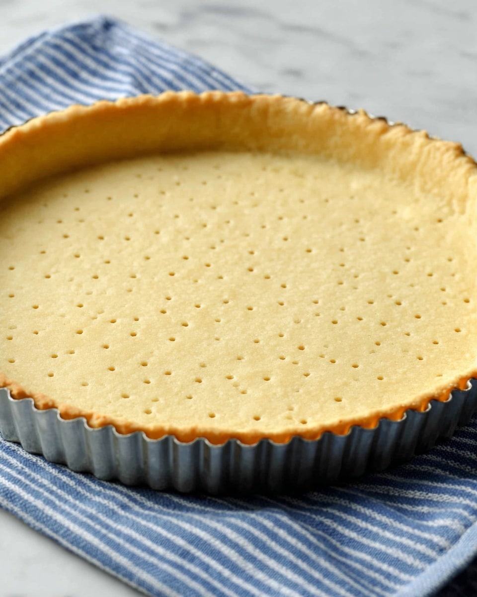 A close-up of a tart shell with one layer of light golden crust, evenly baked with tiny fork holes in the center creating a dotted pattern, resting inside a silver fluted tart pan. The tart pan sits on a folded blue cloth with white stripes, placed on a white marbled surface. The crust edge is crimped with a slightly darker brown color, contrasting the pale, smooth interior. photo taken with an iphone --ar 4:5 --v 7