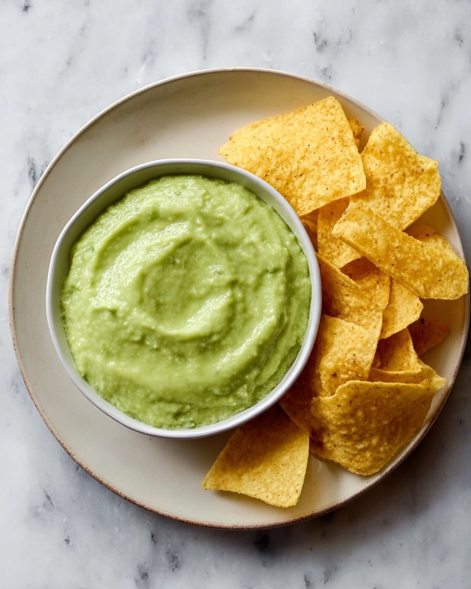 A white plate holds a small bowl filled with smooth, pale green guacamole that has a creamy texture with slight swirls on the surface. On the right side of the bowl, there is a pile of yellowish tortilla chips with a crispy and slightly bumpy texture. The plate is set on a white marbled surface. photo taken with an iphone --ar 4:5 --v 7