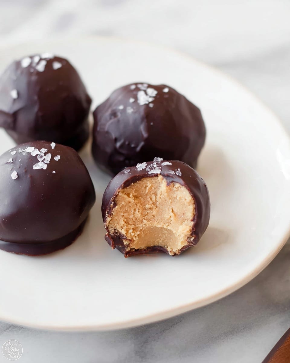 Five round chocolate balls with a smooth dark brown coating are placed together on a white cake stand with a delicate dotted edge. Each chocolate ball is topped with small, shiny flakes of sea salt that add texture and contrast. The chocolate coating looks slightly glossy and has a few small wrinkles, giving a homemade feel. The cake stand is set on a surface with a white marbled texture visible at the edges. photo taken with an iphone --ar 4:5 --v 7