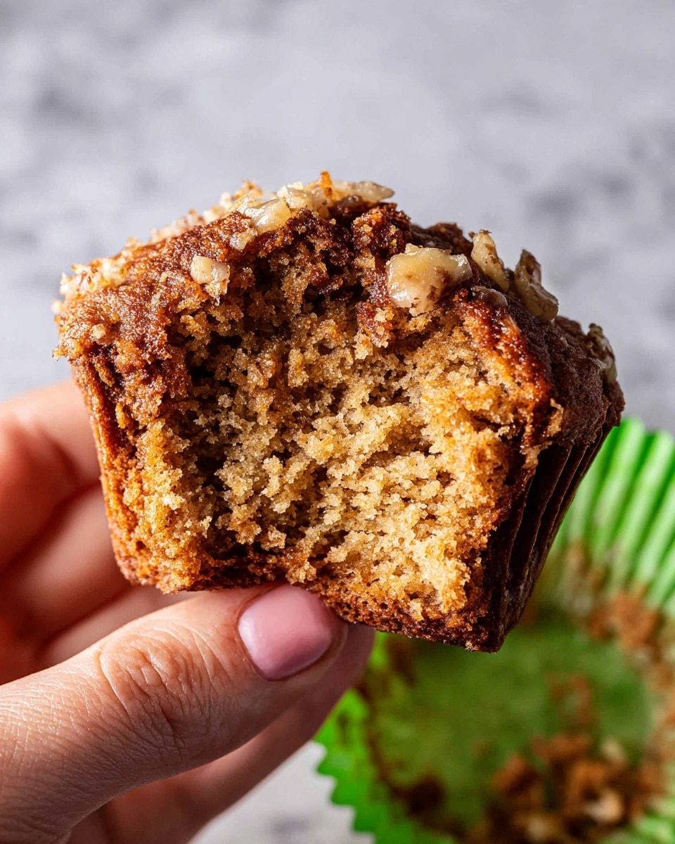 A close-up of a half-eaten brown muffin held by a woman's hand, showing a moist and crumbly texture inside. The muffin's top layer is rough and slightly cracked with small bits of nuts or streusel, while the outer edges are darker and slightly crispy. It sits on a green muffin liner with some brown crumbs scattered on it, and the background is a white marbled texture. photo taken with an iphone --ar 4:5 --v 7