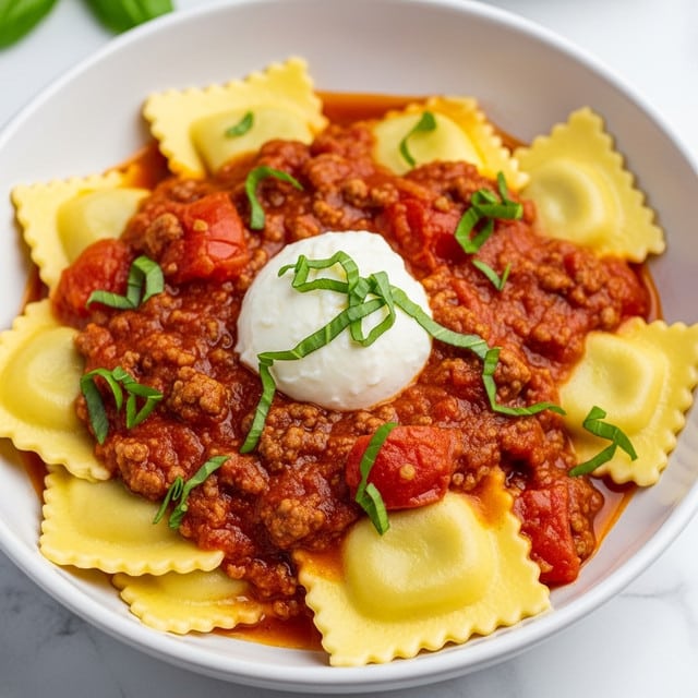 A close-up view of a white bowl filled with ravioli in a thick, rich tomato sauce layered with ground meat and pieces of tomato. The ravioli are pale yellow with ridged edges and are scattered in the sauce. The sauce is mostly orange-red with some oil spots and pieces of ground meat giving a textured look. In the center, there is a soft white dollop of cheese topped with small green basil leaves and fresh chopped basil sprinkled over the dish, adding a contrast of green. The bowl sits on a white marbled surface. Photo taken with an iphone --ar 4:5 --v 7