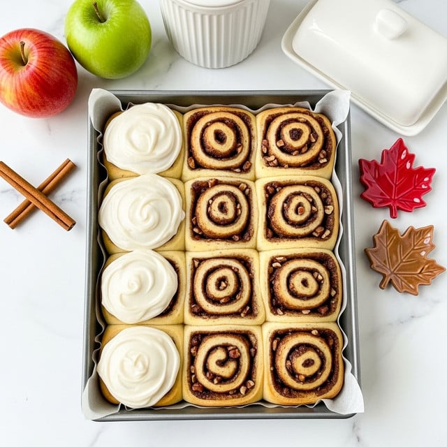 A metal rectangular pan filled with twelve cinnamon rolls arranged in a 3 by 4 grid, some topped with creamy white icing that has a smooth, thick texture, while others show swirls of cinnamon brown with bits of nuts and dark cinnamon filling visible; the rolls have a golden-brown baked edge and soft spiral layers, all resting on parchment paper inside the pan. The background is a white marbled surface with a red apple and green apple on the left, two cinnamon sticks, a white ceramic butter dish, and a white ribbed container behind the pan. On the right side, there are two small red and brown maple leaf-shaped dishes. Photo taken with an iphone --ar 4:5 --v 7