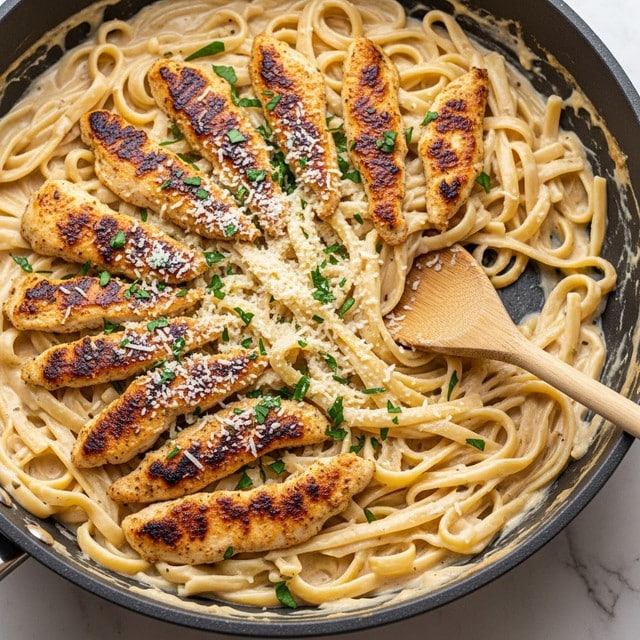 A close-up of a large pan filled with creamy fettuccine pasta coated in a thick, light beige sauce. On top, there are several pieces of golden brown grilled chicken strips evenly spread, their edges slightly charred for texture. The dish is sprinkled with finely grated white cheese and chopped green herbs scattered over the pasta and chicken. A wooden spoon is resting inside the pan, partially submerged in the pasta. The pan is placed on a white marbled surface. photo taken with an iphone --ar 4:5 --v 7