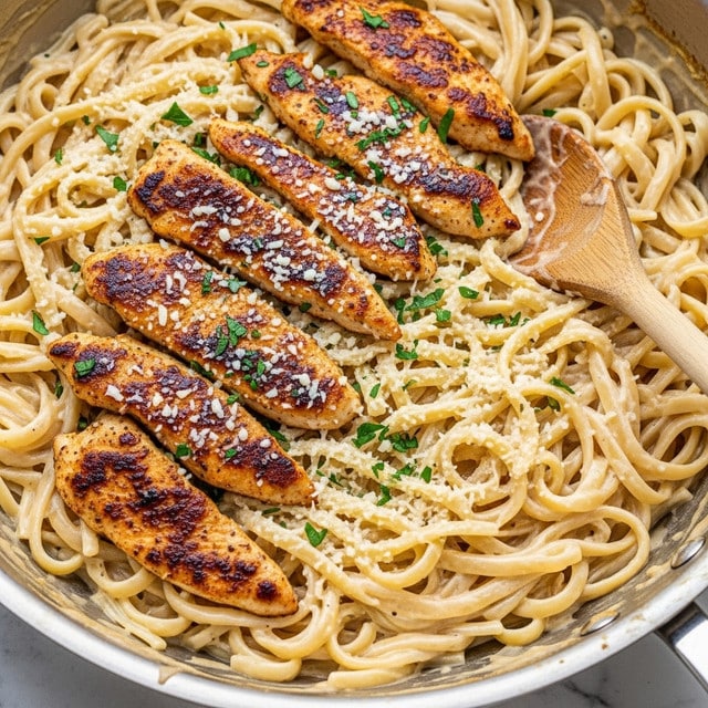 The image shows a close-up of a creamy pasta dish in a stainless steel pan, featuring thick fettuccine noodles coated in a rich, pale yellow sauce. On top of the pasta are several pieces of golden-brown grilled chicken strips, each with a slightly crispy texture. The dish is sprinkled with finely grated white cheese and chopped green herbs, adding color contrast and freshness. A wooden spoon rests on the edge of the pan, partially buried in the pasta, revealing the creamy texture of the sauce. The dish sits on a white marbled surface. photo taken with an iphone --ar 4:5 --v 7
