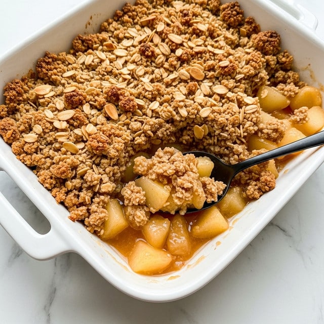 A white rectangular baking dish filled with two main layers: the bottom layer consists of soft, cooked apple pieces in a light brown syrupy sauce, while the top layer is a crumbly, golden brown oat and almond topping with a slightly rough texture. A dark spoon is inserted into one corner, scooping up some of the apple filling and crunchy topping, highlighting the contrast between the juicy fruit and the crisp crumble. The dish rests on a white marbled surface. photo taken with an iphone --ar 4:5 --v 7
