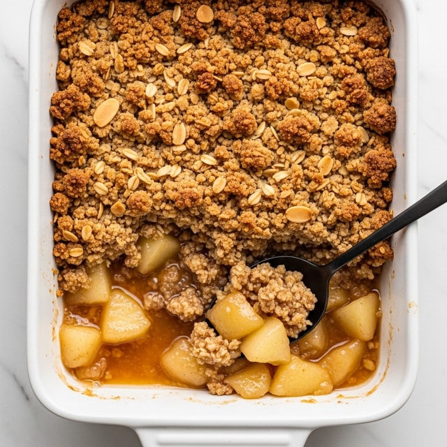 A close-up view of a white rectangular baking dish filled with baked apple crumble, showing a thick top layer of golden-brown crumbly texture mixed with sliced almonds, beneath which soft, cooked chunks of light brown apples are visible, with a silver spoon scooping some apples from the dish, all placed on a white marbled surface. photo taken with an iphone --ar 4:5 --v 7