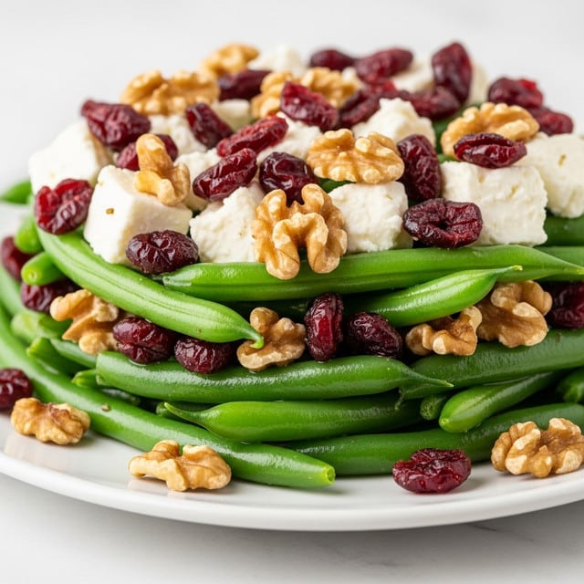 The close-up view shows a layered green bean salad on a white plate. The bottom layer is bright, fresh green beans with a smooth and slightly shiny texture. Scattered on top of the green beans are chunks of white, crumbly cheese, adding a soft contrast. There are deep red dried cranberries spread evenly among the green beans, adding color and texture. Whole walnut pieces with a light brown, rough surface are placed throughout the salad. The whole dish sits on a white marbled surface, highlighting the colors and textures of the salad. photo taken with an iphone --ar 4:5 --v 7