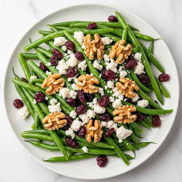 The image shows a fresh green bean salad served on a white plate. The salad has several layers: the base layer is bright green beans that look cooked but still crisp with a shiny texture. On top of this are scattered white crumbly cheese pieces and dark red dried cranberries, adding color contrast. Whole walnut halves are also placed evenly over the salad, adding a brown crunchy element. The plate is set on a white marbled surface, giving a clean fresh look. photo taken with an iphone --ar 4:5 --v 7