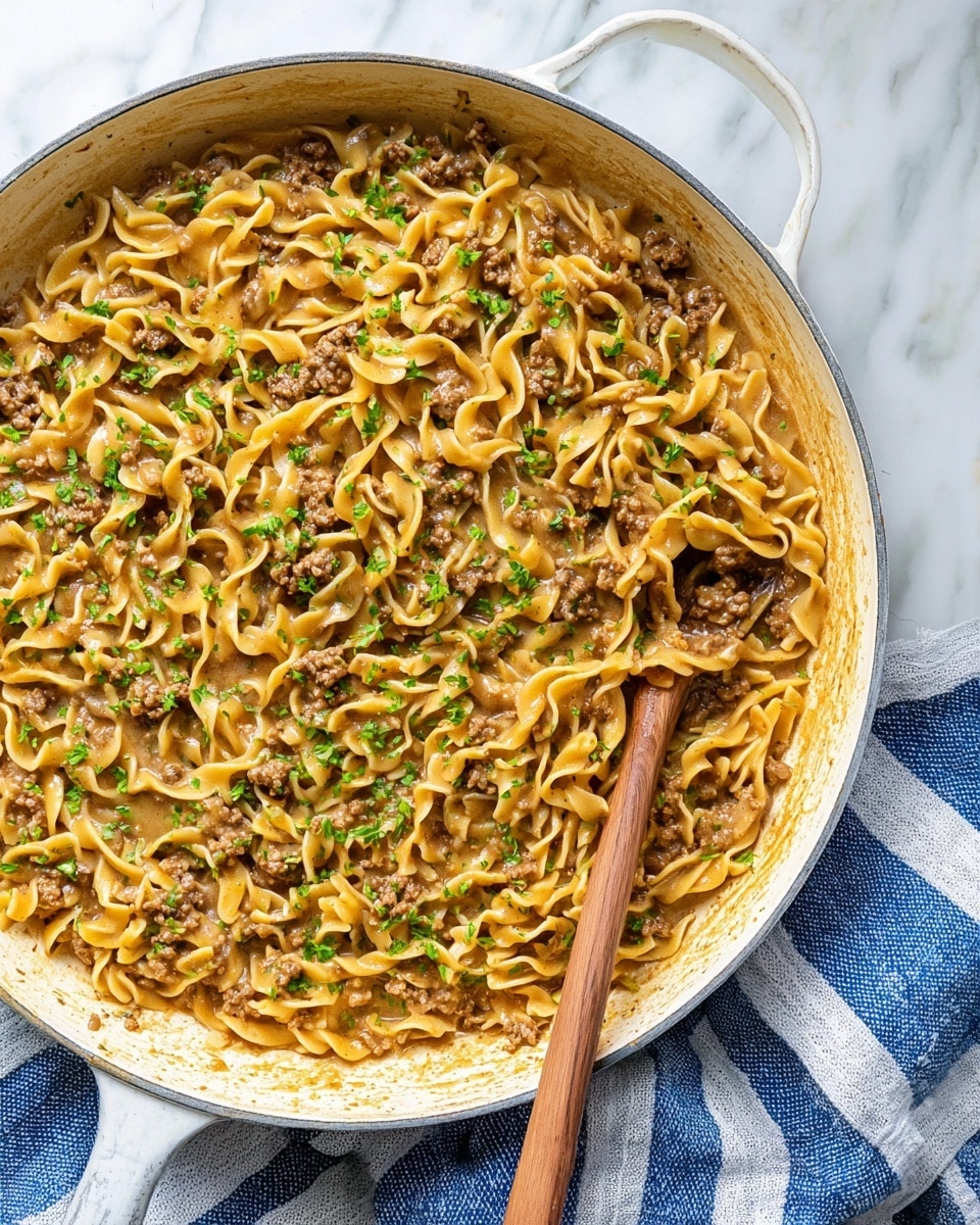 A white pan filled with a hearty dish made of three main layers: the bottom layer is a light brown sauce with a slightly creamy texture, the middle layer consists of cooked ground beef pieces scattered evenly, and the top layer features golden yellow twisted egg noodles mixed throughout. Some small bits of cooked onions are also visible, adding a pale translucent color. The dish is sprinkled with finely chopped green parsley for a fresh look. A wooden spoon is resting inside the pan, covered with the mixture. The pan sits on a white marbled surface, next to a blue and white striped cloth. Photo taken with an iphone --ar 4:5 --v 7