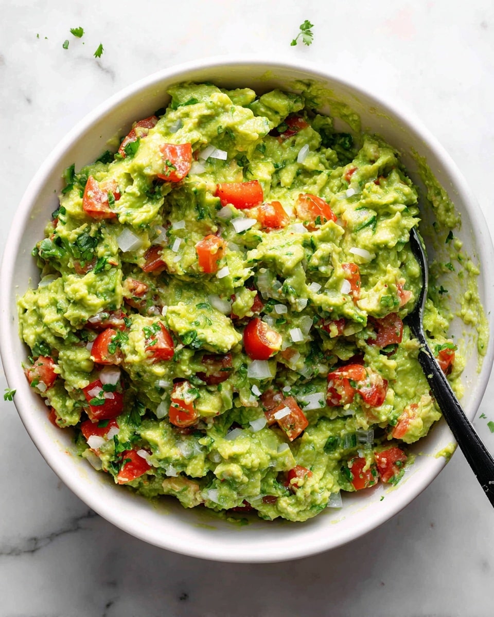 A close-up view of a white bowl filled with chunky guacamole showing three main layers of colors and textures: bright green mashed avocado forming the thick base with its smooth creamy texture broken by some larger pieces, small red tomato chunks evenly scattered throughout adding pops of color, and finely diced white onions mixed in for contrast. There are also bits of dark green cilantro leaves spread across the mix. A black spoon sits inside the bowl on the right side, partially submerged in the guacamole. The bowl is set on a white marbled surface with faint gray veins visible. Photo taken with an iphone --ar 4:5 --v 7