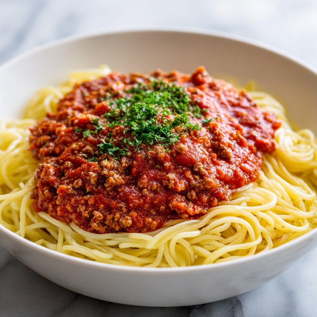A white bowl holds a serving of spaghetti pasta, with smooth, yellow strands forming the base layer. On top, there is a thick layer of rich, dark red meat sauce with small chunks of ground meat, spread evenly over the noodles. The sauce is garnished with small green parsley bits sprinkled in the center. The background features a white marbled texture. Photo taken with an iphone --ar 4:5 --v 7