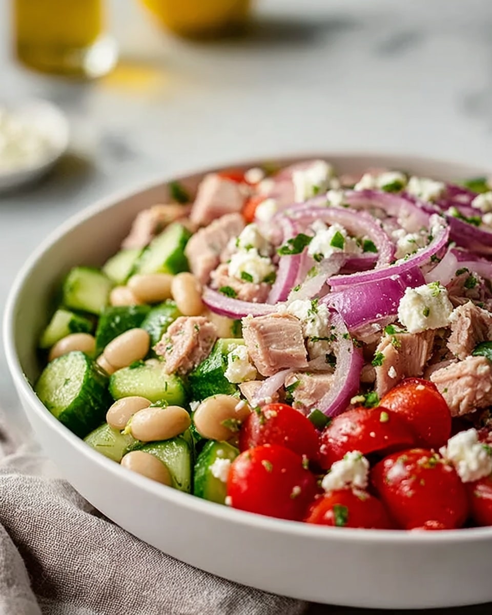 The image shows a bowl of layered salad on a white marbled surface. The base layer is a mix of white beans and small green cucumber slices, topped with bright red cherry tomatoes cut in halves. Scattered throughout are chunks of light pink tuna and crumbled white feta cheese. Thin slices of purple red onion are spread across the top, and finely chopped green herbs are sprinkled all over, adding a fresh green touch. The bowl itself is white, rounded with a smooth texture. Photo taken with an iphone --ar 4:5 --v 7