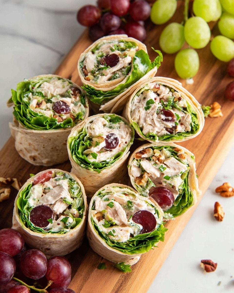 Several tortilla roll-up slices are arranged closely on a wooden board. Each roll-up has a light beige outer tortilla layer wrapped around layers of bright green leafy lettuce, creamy white chicken salad mixed with small pieces of nuts, and halved red grapes showing their juicy, glossy interior. Small green celery pieces and finely chopped herbs are scattered inside and on top, adding fresh texture and color. Around the board, whole red grapes, celery sticks, and nut pieces rest on a white marbled surface. photo taken with an iphone --ar 4:5 --v 7