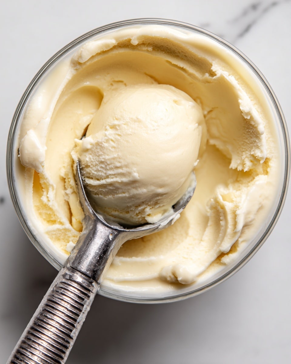 A top view of a round clear container filled with smooth, creamy pale yellow ice cream, with a shiny metal ice cream scoop resting inside. The scoop has a ridged handle and holds a round, glossy scoop of the same pale yellow ice cream, slightly raised above the surface. The inside edges of the container show soft scooping marks with a fluffy texture, sitting on a white marbled surface. photo taken with an iphone --ar 4:5 --v 7