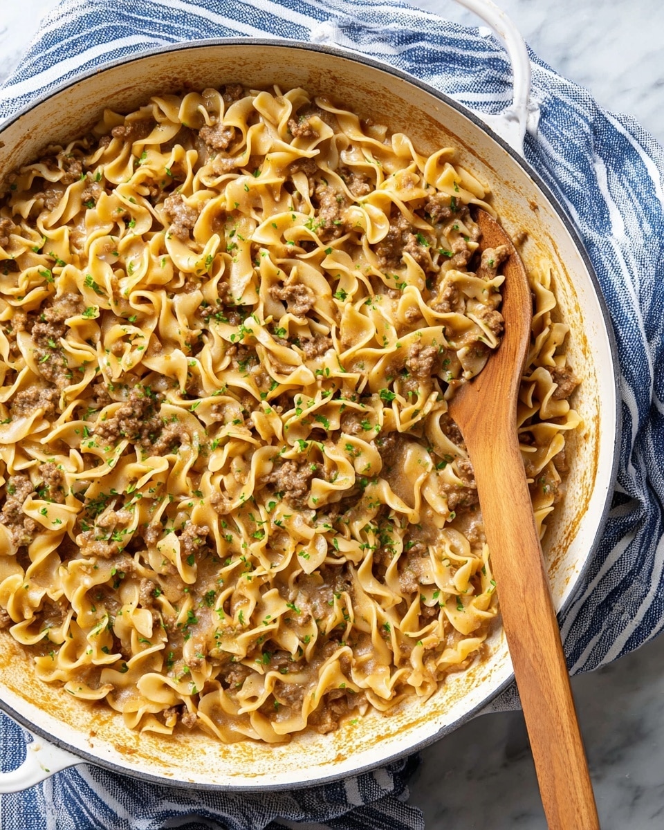 A large white pan filled with a one-layer dish of twisted egg noodles cooked with small ground meat bits and chunks of soft white onion, covered in a light brown gravy sauce that edges the pan with a slightly darker toasted residue; the noodles are sprinkled with small, bright green parsley pieces and a wooden spoon with a light brown handle rests on the right side inside the pan; the pan sits on a white marbled surface with a blue and white striped cloth partially underneath. photo taken with an iphone --ar 4:5 --v 7