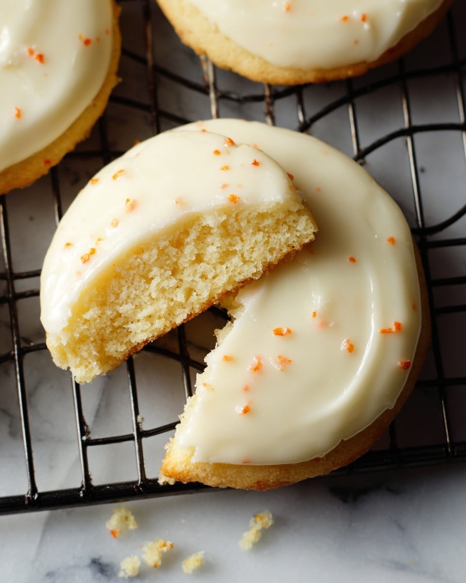 A single round cookie with a smooth, creamy white icing on top, showing tiny orange specks inside the icing. The cookie has a bite taken out of its left side, revealing a soft, pale yellow inside. It rests on a black metal cooling rack, with some small crumbs on the rack near the cookie. The background is a white marbled texture. photo taken with an iphone --ar 4:5 --v 7