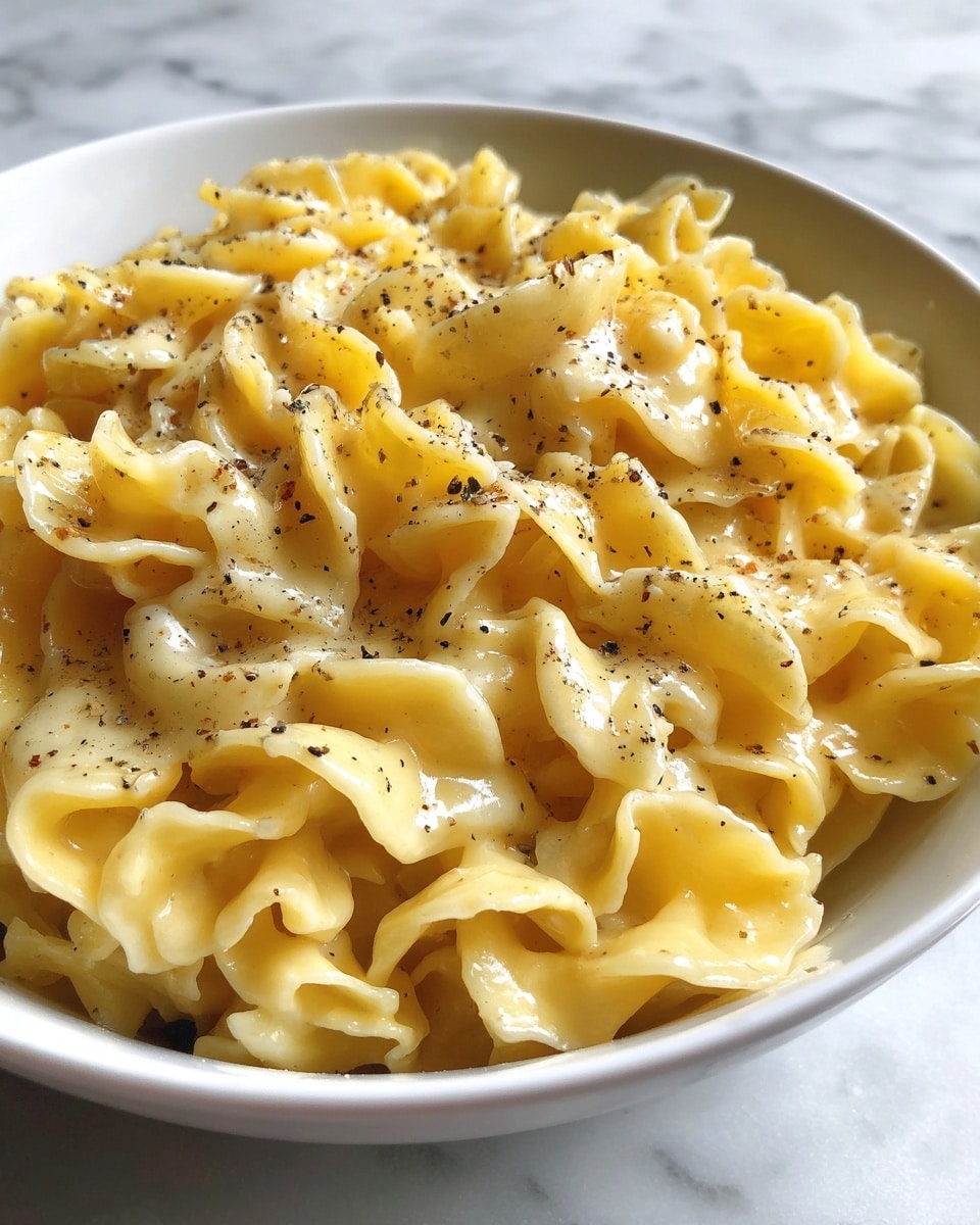 A close-up view of creamy macaroni and cheese in a white bowl, showing thick, wavy pasta coated in smooth, shiny, light yellow cheese sauce with small black pepper specks sprinkled on top. The edges of the pasta have a slightly ruffled texture and the cheese looks rich and velvety, filling the bowl generously. The background is a white marbled surface. photo taken with an iphone --ar 4:5 --v 7