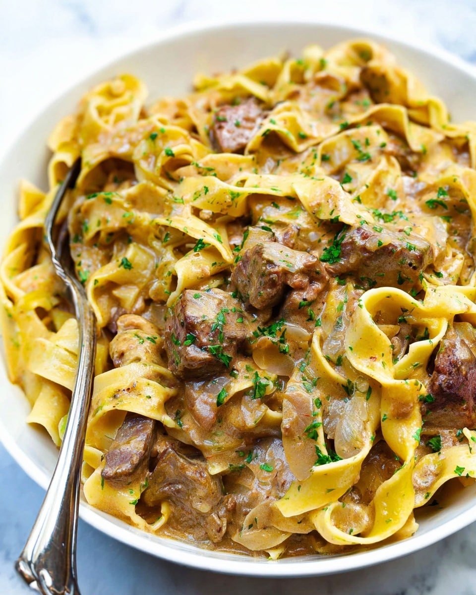 A close-up view of a white bowl filled with a creamy pasta dish showing thick, yellow noodles layered and twisted throughout. Among the pasta are medium-sized chunks of brownish-gray meat and small pieces of creamy off-white mushrooms. The dish is lightly sprinkled with small green herb bits, and a silver fork is partially buried in the pasta, all placed on a white marbled surface. The overall texture looks soft and saucy, with a warm, inviting color palette. photo taken with an iphone --ar 4:5 --v 7