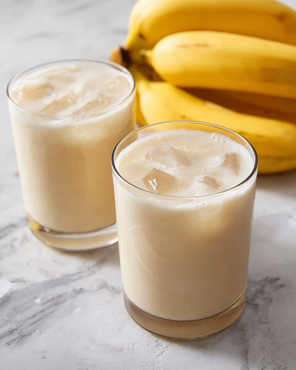 Two clear glasses filled with a creamy light beige banana smoothie are placed on a white marbled surface. The smoothie has a smooth, thick texture with a few gentle swirls and ice cubes visible inside the drink. In the background, a bunch of ripe yellow bananas sits, adding a fresh and natural touch. The overall look is fresh, smooth, and inviting, showing the creamy layers and cold ice in the glass. Photo taken with an iphone --ar 4:5 --v 7