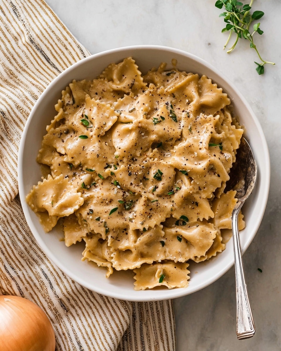 A white bowl filled with two layers of wide, ruffled-edge pasta coated in a creamy, light beige sauce. The pasta is evenly covered, with visible seasoning of black pepper and small green herb leaves sprinkled on top. A silver fork rests on the right edge of the bowl, partly submerged in the pasta. The bowl sits on a white marbled surface, with a peeled onion at the bottom left corner and a small sprig of herbs near the top right. A striped cloth is softly placed behind the bowl. photo taken with an iphone --ar 4:5 --v 7