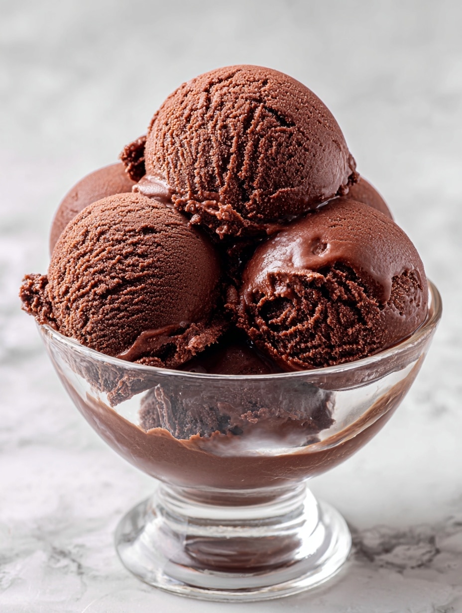 The image shows four smooth and creamy scoops of rich brown chocolate ice cream stacked in a clear glass cup. Each scoop has a slightly rough texture on the edges with frosty ice crystals visible, adding a fresh, cold feel. The scoops fill the cup tightly, with some small chocolate ice cream pieces sticking out from the sides. The cup sits on a white marbled background, enhancing the deep brown color of the ice cream. photo taken with an iphone --ar 4:5 --v 7