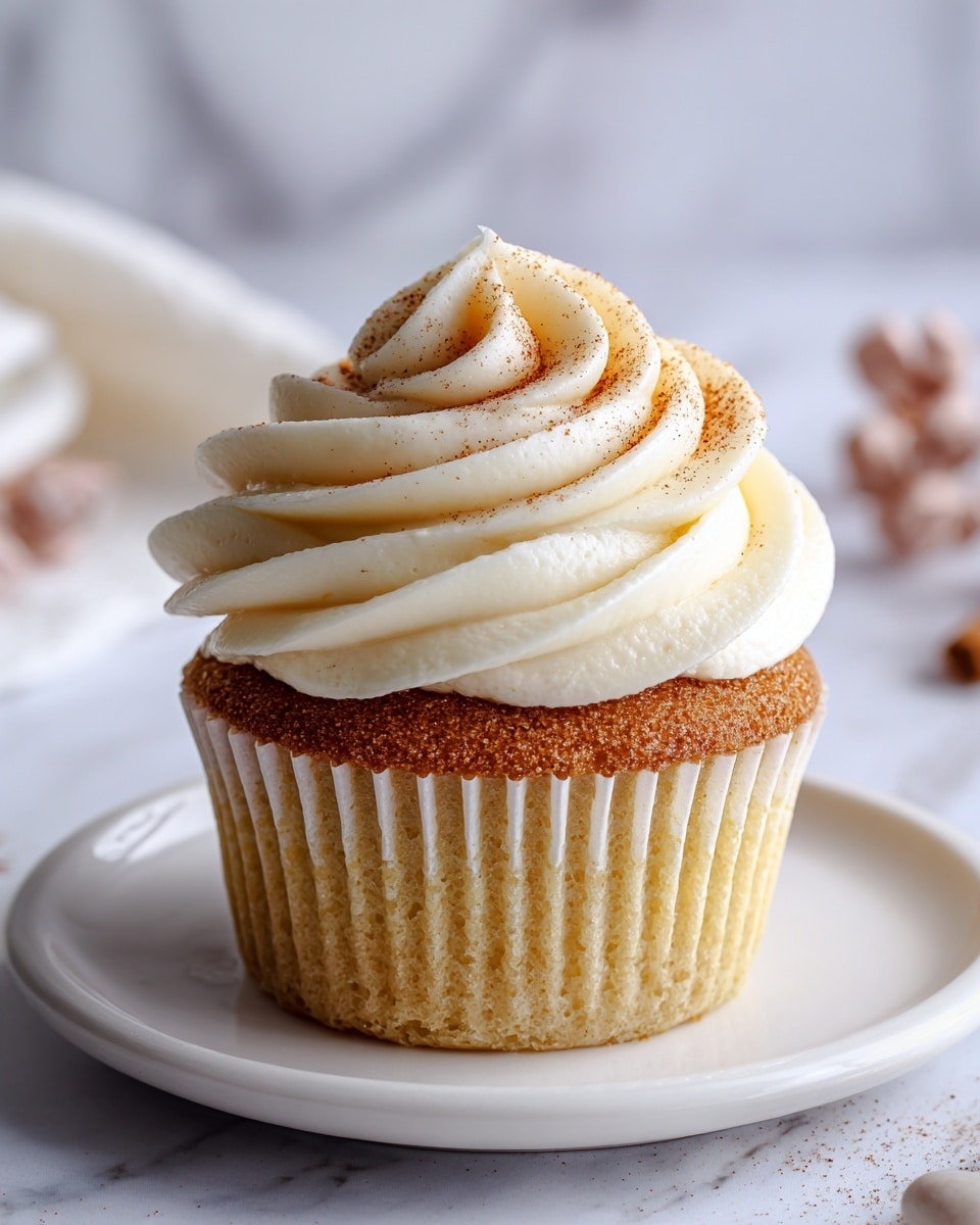A single cupcake sits centered on a white plate with a black rim, placed on a white marbled surface. The cupcake has three main layers: the base is a golden brown cake with a soft texture, followed by a thick swirl of smooth white frosting that is styled in a flower shape, and topped with a smaller spiral of light brown cinnamon-flavored frosting dusted with fine cinnamon powder. The contrast between the warm cake color and the creamy white frosting creates a visually appealing treat. photo taken with an iphone --ar 4:5 --v 7