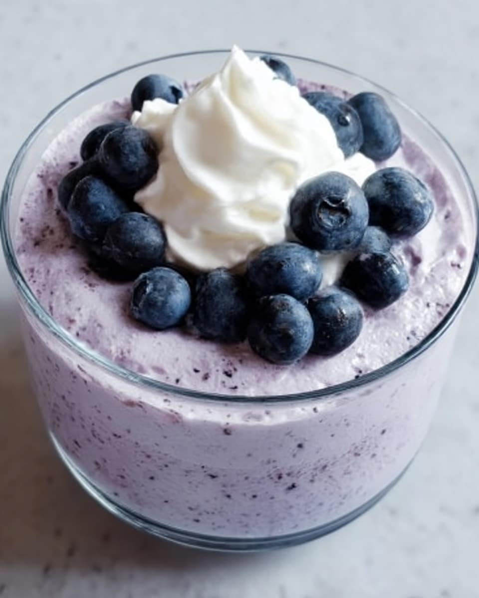 A clear glass bowl filled with a light purple mousse, smooth and creamy in texture, topped with a dollop of white whipped cream placed in the center. On top of the whipped cream are several whole dark blue blueberries scattered evenly. The bowl sits on a white marbled surface. Photo taken with an iphone --ar 4:5 --v 7