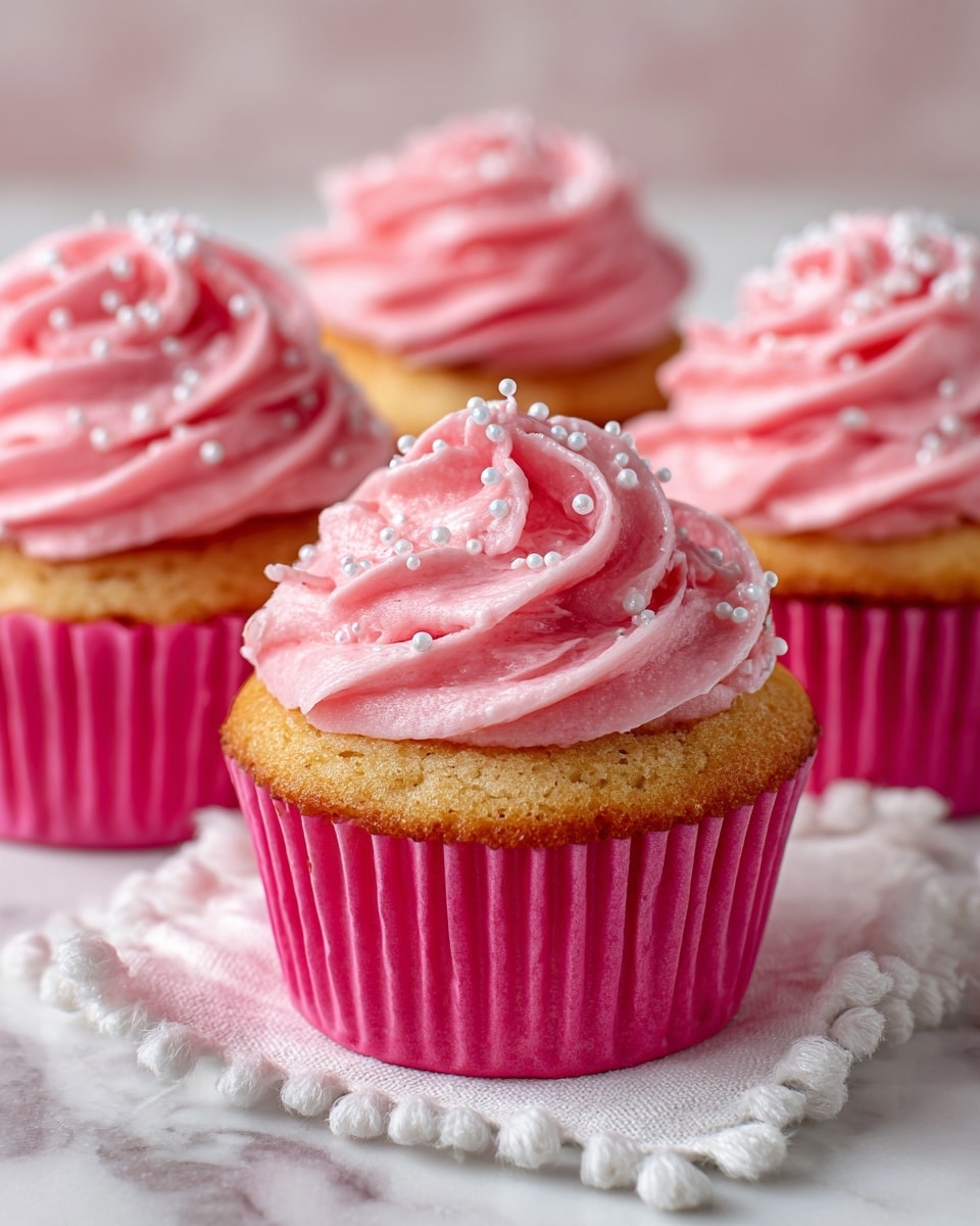 The image shows four cupcakes arranged closely on a soft white cloth with small pom-poms on the edges, placed over a white marbled textured surface. Each cupcake has a bright pink wrapper and a swirled, tall frosting on top in a smooth, creamy pink color, decorated with a few small white round sprinkles. The cupcakes' cake layer at the bottom is light golden brown. They are positioned in a way that three are in the background while one cupcake with the most visible frosting is centered in the front. Photo taken with an iphone --ar 4:5 --v 7