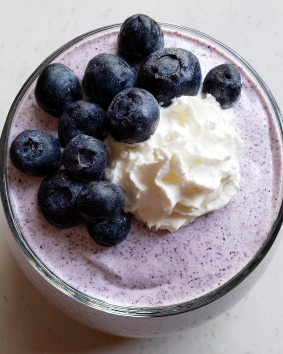 A clear glass bowl contains a creamy light purple mousse with small dark purple specks mixed in. On top, there is a dollop of smooth white whipped cream placed slightly off-center. Surrounding and sitting on the whipped cream are fresh, whole blueberries with a deep blue color and a natural matte texture. The bowl rests on a white marbled surface. Photo taken with an iphone --ar 4:5 --v 7