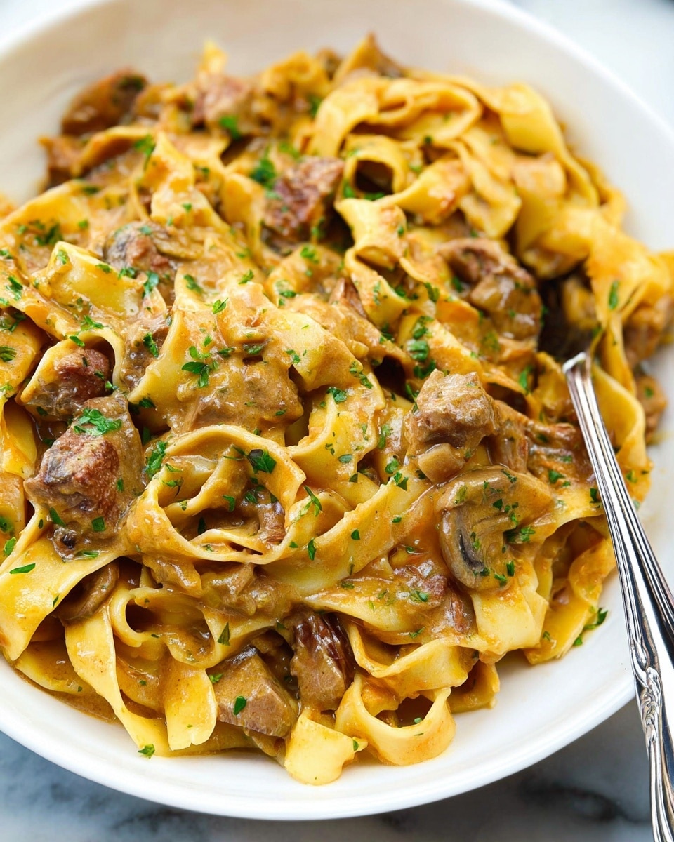 This image shows a close-up of a white bowl filled with wide, flat, yellow pasta noodles that are soft and curly, mixed with chunks of brown meat and small pieces of light-colored mushrooms. The pasta and meat are covered in a thick, creamy brownish sauce that looks rich and a bit glossy. Bright green chopped herbs are sprinkled on top, adding a fresh touch to the dish. A silver fork is partially buried in the food, with the handle sticking out toward the bottom left of the bowl. The whole scene is set against a white marbled surface. photo taken with an iphone --ar 4:5 --v 7