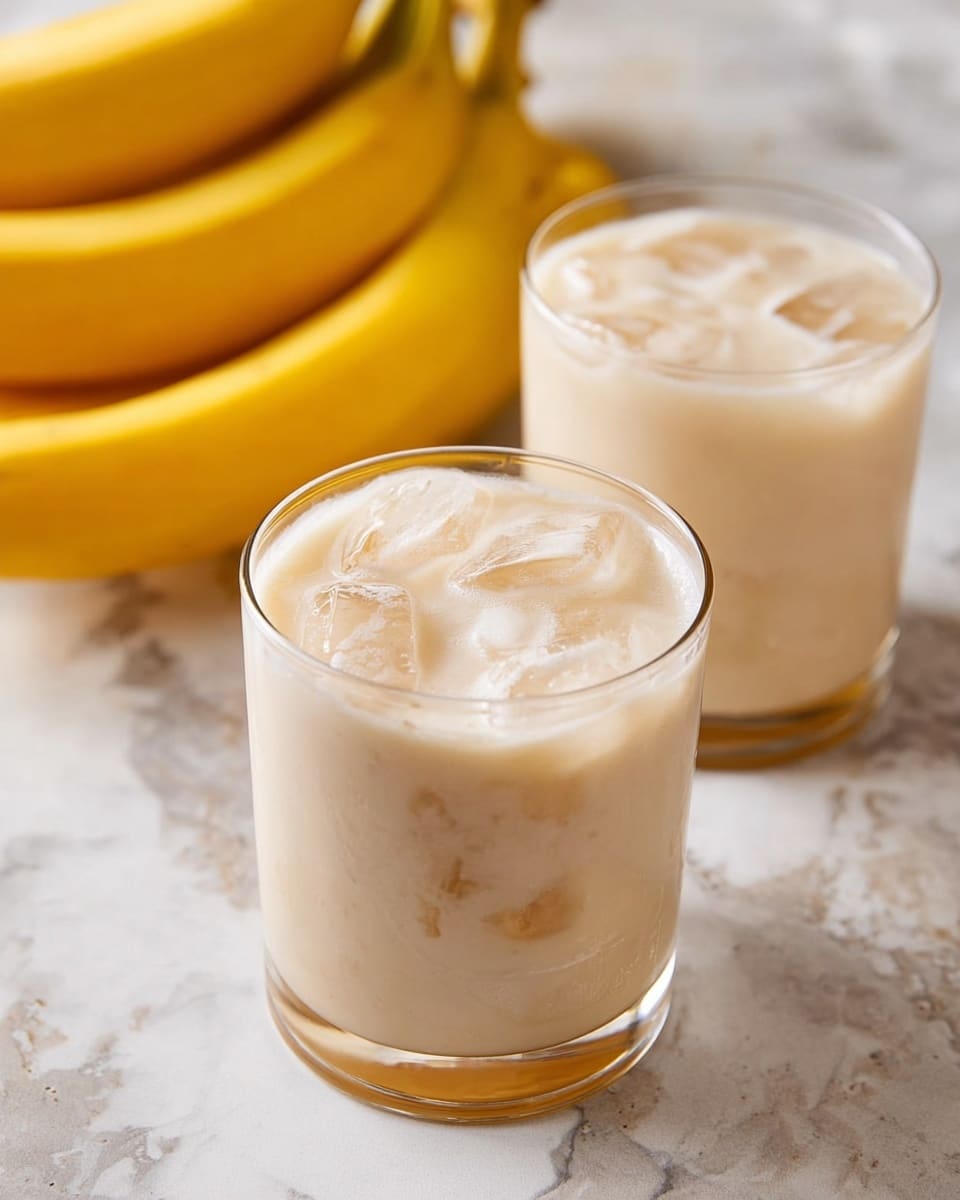 Two clear glasses filled with a creamy, light beige banana smoothie are placed on a surface with a white marbled texture. Each glass shows a layer of frothy foam on top, with visible ice cubes giving a translucent effect within the smooth mix. Behind the glasses, a bunch of ripe yellow bananas adds a pop of bright color and hints at the drink’s flavor. The overall look is fresh, with smooth creamy textures and soft natural light. photo taken with an iphone --ar 4:5 --v 7