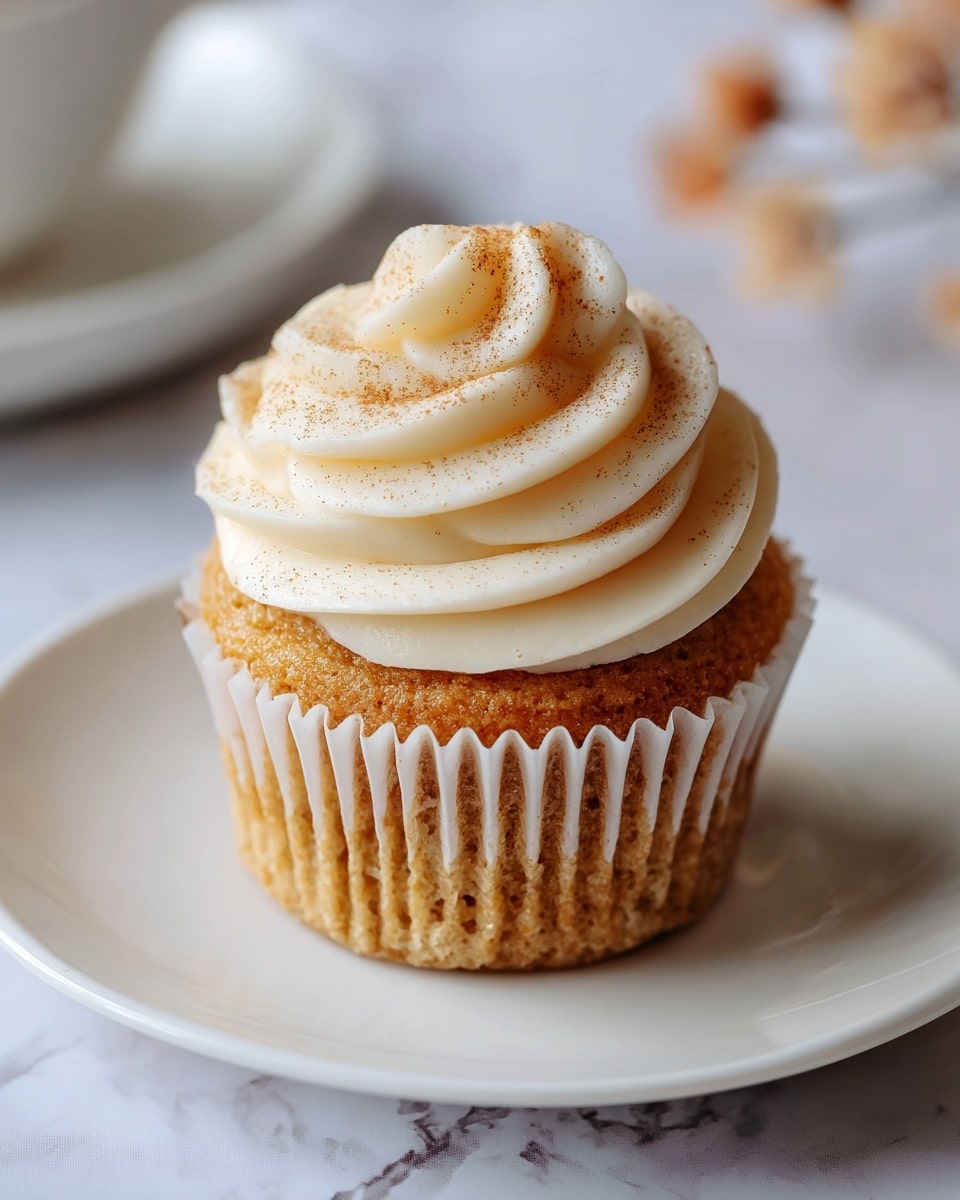 A single cupcake is centered on a white cupcake liner with ridged edges, placed on a white plate with a simple design. The cupcake has two main layers: the base is a golden brown cake with a soft, moist texture, and on top is a thick swirl of creamy white frosting with smooth and slightly ridged edges. In the middle of the frosting swirl is a smaller spiral of light brown icing or cinnamon dusting, adding a touch of color and texture contrast. The plate rests on a white marbled surface with soft, faint veins, and a few small, out-of-focus decorative elements are visible around the edges. photo taken with an iphone --ar 4:5 --v 7