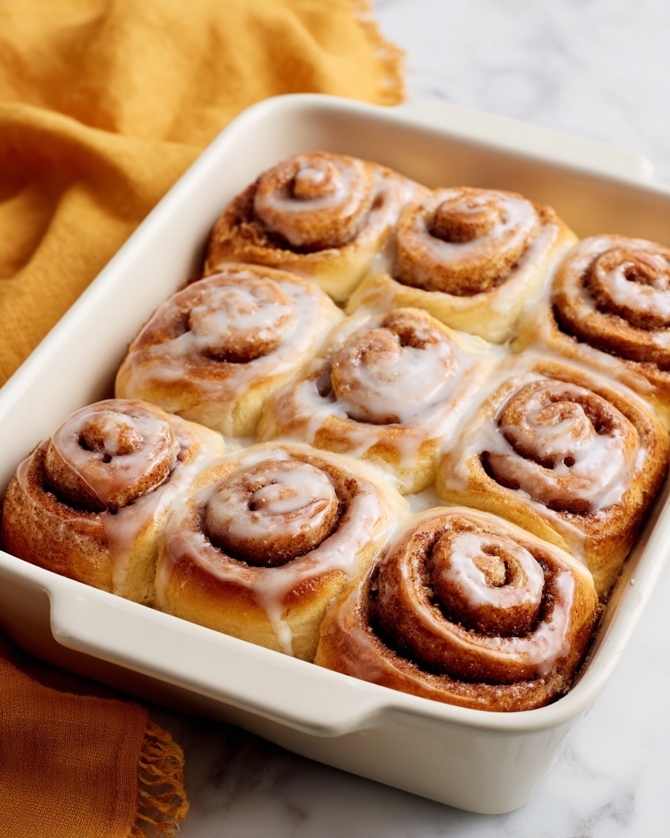 A white baking dish holds six golden brown cinnamon rolls tightly packed in two rows of three each. Each cinnamon roll shows clear spiral layers: the outer layer is soft and light golden, with a thick, sticky cinnamon-sugar layer in the middle, dark brown and slightly crumbly. A white glaze lightly covers the top of each cinnamon roll, dripping softly down the sides. The dish is placed on a white marbled surface, with a soft, blurred mustard yellow cloth in the background. photo taken with an iphone --ar 4:5 --v 7