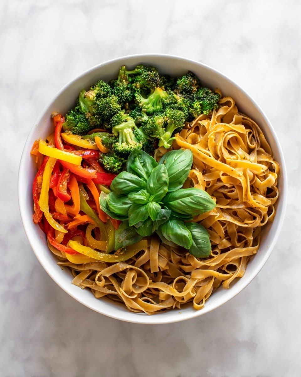 The dish shows a bowl filled with flat, wide noodles layered at the bottom with a smooth, light beige color and slightly glossy texture. On top, there are bright green broccoli florets with a rough, leafy texture, orange and red bell pepper slices that are smooth and curved, and yellow strips that add brightness. Fresh green basil leaves with a slight sheen sit in the center as a garnish. The bowl is white and the dish rests on a white marbled surface. photo taken with an iphone --ar 4:5 --v 7