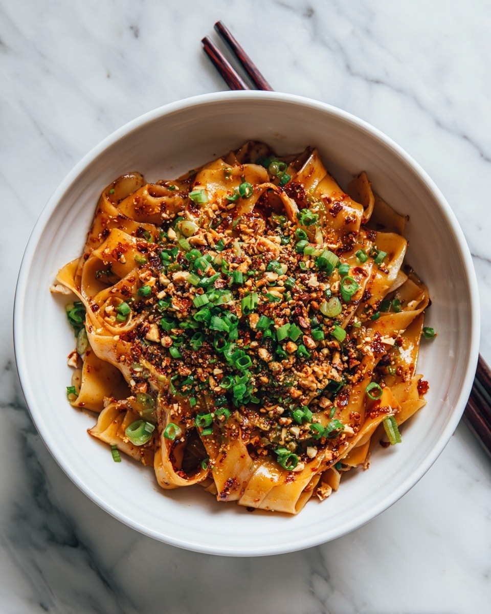 The image shows a bowl filled with a dish of wide, flat noodles tossed in a reddish-brown sauce. On top, there is a thick layer of chopped green onions and crushed nuts. The noodles look soft and slightly glossy, mixed with small bits of red chili flakes. The bowl is white and sits on a white marbled surface. A woman's hand is holding black chopsticks resting on the edge of the bowl. photo taken with an iphone --ar 4:5 --v 7