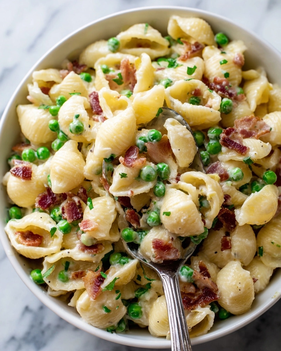 A close-up image of a white bowl filled with a creamy pasta salad featuring small shell pasta as the base layer, pale yellow in color with a smooth texture. Mixed within the pasta are bright green peas scattered evenly, adding a pop of color and round texture. Small pieces of crispy, brown bacon are spread throughout the dish, providing contrast and a rustic look. The creamy sauce lightly coats all ingredients, giving a glossy appearance. A silver spoon scoops a portion of the salad up, showing a mix of all elements. The background is a white marbled surface. Photo taken with an iphone --ar 4:5 --v 7