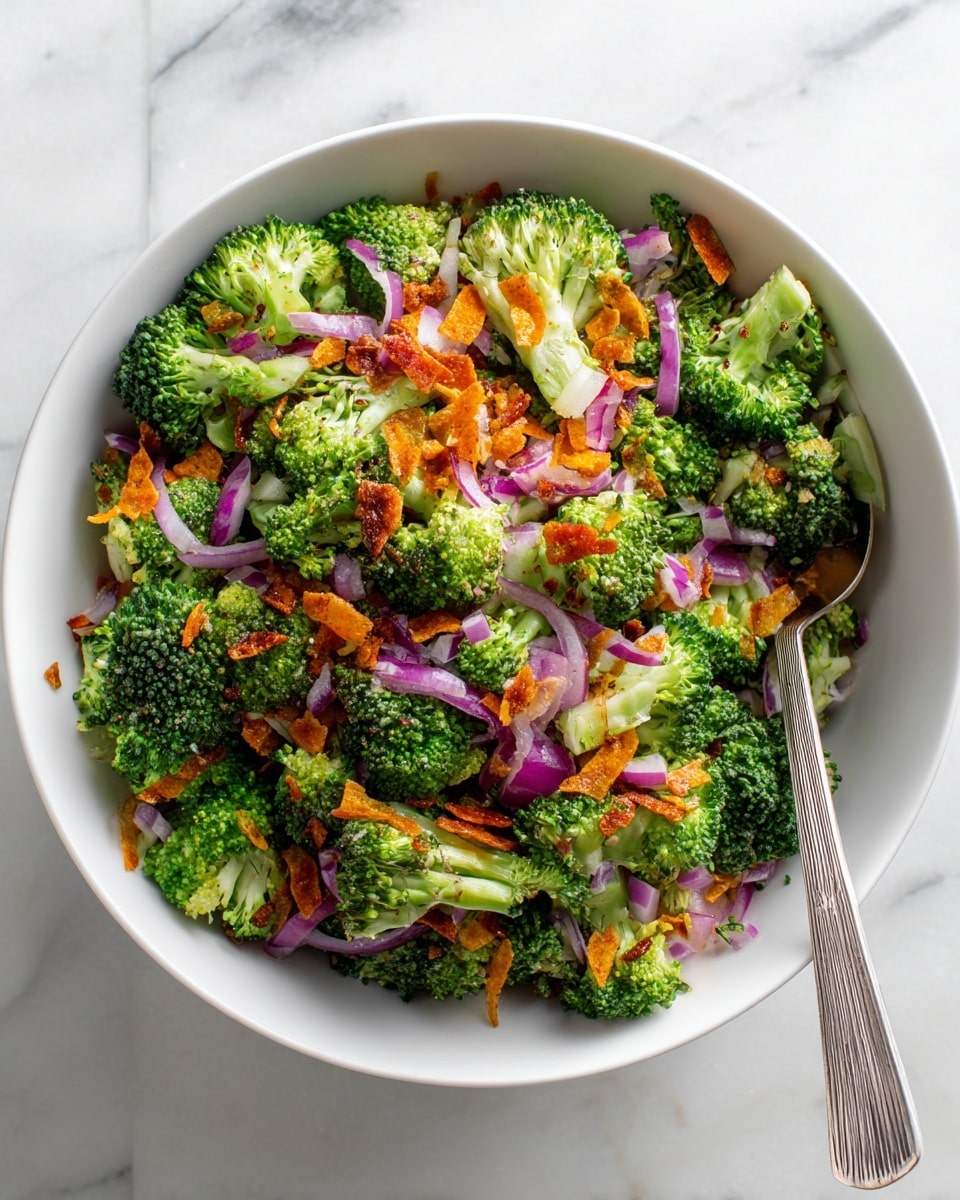 A white bowl filled with a fresh broccoli salad showing three main layers: bright green broccoli florets as the base, small chunks of purple-red onion and orange carrot slices scattered evenly on top, and thin crispy golden-brown chip-like pieces sprinkled over everything, creating a vibrant mix of textures and colors; a silver spoon is placed inside the bowl on the right side, all set on a white marbled surface photo taken with an iphone --ar 4:5 --v 7
